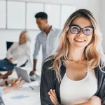 Eine fröhliche junge Frau mit Brille steht im Vordergrund, die Arme verschränkt und ein selbstbewusstes Lächeln auf den Lippen, während im Hintergrund ihre Kollegen konzentriert an einem Projekt arbeiten. Sie verkörpert die erfolgreichen Bewerber:innen, die von unseren Leistungen profitiert haben und nun Teil eines dynamischen Teams sind.