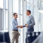 Zwei Geschäftsmänner in legerer Bürokleidung geben sich die Hand in einem modernen Büro mit Glasfronten, was erfolgreiche Verhandlungen oder eine Einigung zwischen Fachkräften für Medizintechnik darstellt. Sie lächeln sich gegenseitig zu, was auf eine positive Geschäftsbeziehung hindeutet und die Wichtigkeit von professionellen Partnerschaften in der Medizintechnikbranche unterstreicht.