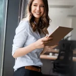 Eine lächelnde Frau in Business-Kleidung steht neben einem Fenster und hält ein Tablet in den Händen. Sie trägt eine helle Bluse und schwarze Hosen mit einem geflochtenen Gürtel. Im Hintergrund ist ein modernes Büro zu sehen.