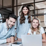glückliche junge Geschäftskollegen, die im Büro mit Geräten arbeiten und in die Kamera lächeln