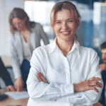 Eine Frau im Vordergrund, die selbstbewusst in die Kamera lächelt, während im Hintergrund ein Team von Kollegen in einer Besprechung arbeitet, die sich auf Steuerberatung und Finanzwesen konzentrieren.