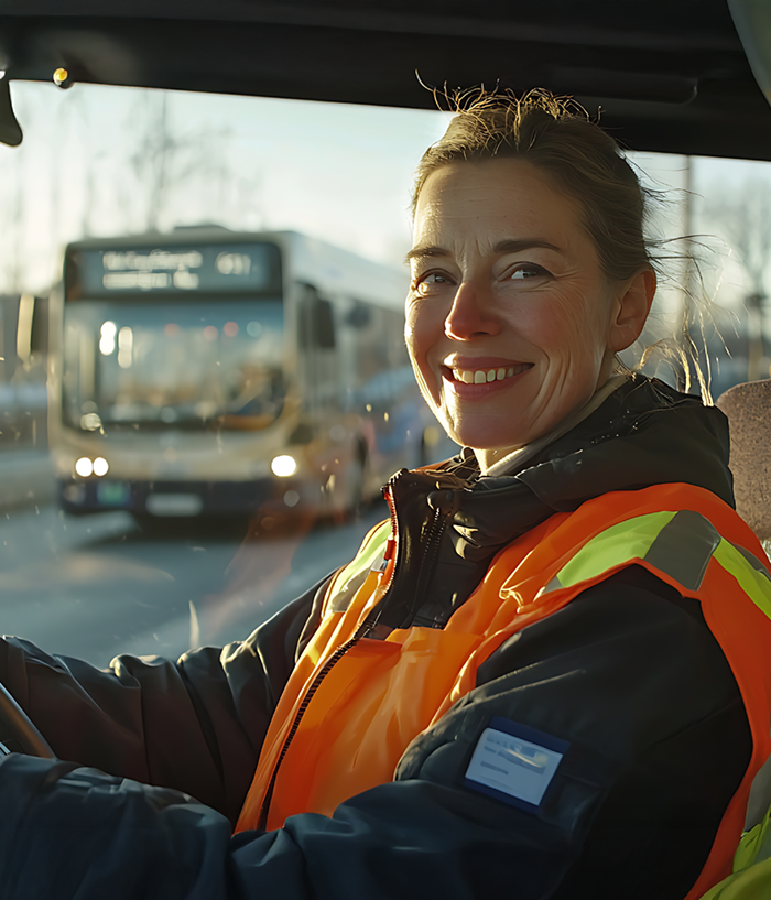 Fahrerin in Warnweste sitzt am Steuer eines Fahrzeugs im öffentlichen Nahverkehr und lächelt in die Kamera. Im Hintergrund fährt ein Bus vorbei. Arbeitssituation aus dem Umfeld von Jobs in öffentlichen Verkehrsbetrieben.