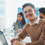 Mitarbeiter in einem modernen Büro sitzt am Schreibtisch und lächelt in die Kamera. Im Hintergrund arbeiten weitere Kolleginnen und Kollegen in einem offenen Office-Bereich. Arbeitssituation aus dem Umfeld kaufmännischer Berufe.