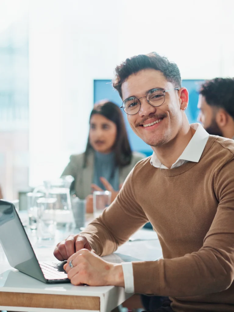 Mitarbeiter in einem modernen Büro sitzt am Schreibtisch und lächelt in die Kamera. Im Hintergrund arbeiten weitere Kolleginnen und Kollegen in einem offenen Office-Bereich. Arbeitssituation aus dem Umfeld kaufmännischer Berufe.