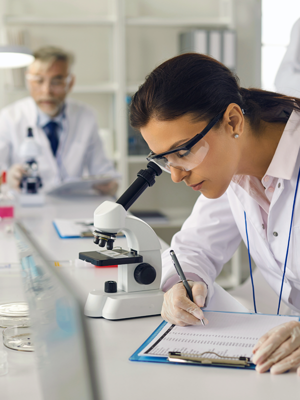 Mitarbeiterin in der Pharmaindustrie arbeitet im Labor, untersucht eine Probe unter dem Mikroskop und macht Notizen. Im Hintergrund arbeitet ein weiterer Kollege.