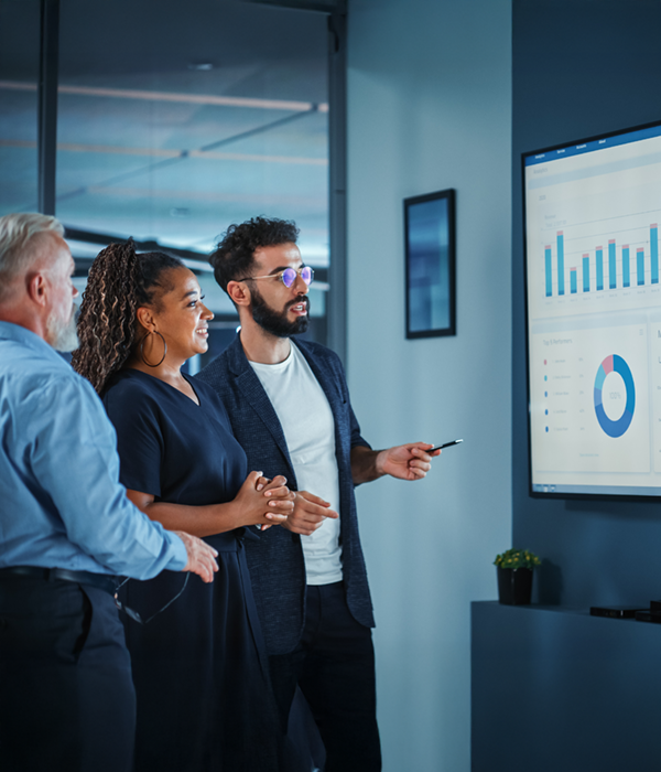 Drei Mitarbeitende stehen in einem modernen Büro vor einem großen Bildschirm mit Diagrammen und Statistiken. Ein Mitarbeiter präsentiert die Daten, während die anderen aufmerksam zuhören. Szene aus dem Arbeitsumfeld kaufmännischer Berufe.