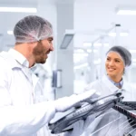 Zwei Mitarbeitende in Reinraumkleidung arbeiten gemeinsam an Bauteilen in einer modernen Produktionsumgebung der Halbleiterherstellung.