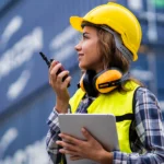 Mitarbeiterin in Warnweste und Schutzhelm steht vor Containern, hält ein Tablet und spricht über ein Funkgerät. Arbeitssituation aus dem Umfeld von Jobs in der Logistik.