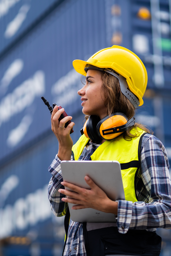 Mitarbeiterin in Warnweste und Schutzhelm steht vor Containern, hält ein Tablet und spricht über ein Funkgerät. Arbeitssituation aus dem Umfeld von Jobs in der Logistik.