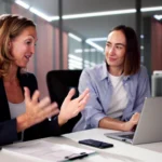 Berufserfahrene Führungskraft im Gespräch mit einer jüngeren Kollegin an einem Laptoparbeitsplatz. Beide sitzen in einem modernen Büro und tauschen sich motiviert über Projekte und Aufgaben aus.