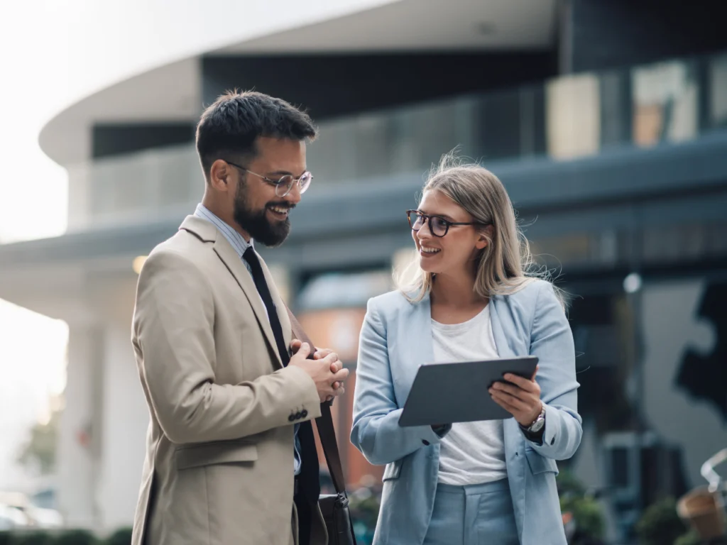 Junge Berufseinsteigerin im Gespräch mit einem Kollegen im Business-Outfit. Beide stehen vor einem modernen Bürogebäude und tauschen sich freundlich über berufliche Aufgaben und Einstiegsmöglichkeiten aus.