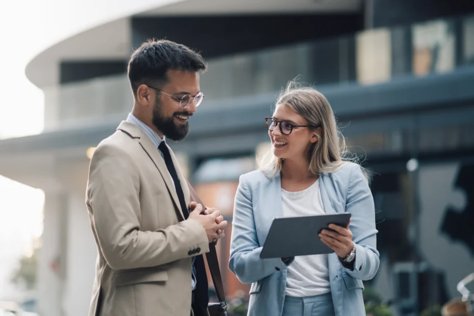 Junge Berufseinsteigerin im Gespräch mit einem Kollegen im Business-Outfit. Beide stehen vor einem modernen Bürogebäude und tauschen sich freundlich über berufliche Aufgaben und Einstiegsmöglichkeiten aus.