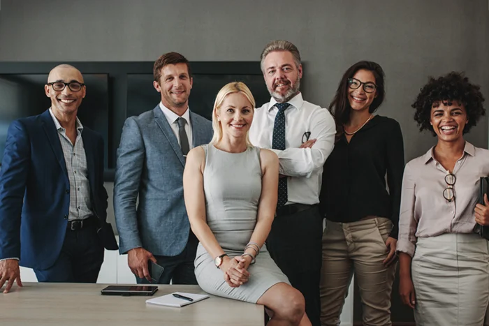 Gruppenfoto eines professionellen Führungsteams in einem modernen Büro: fünf Frauen und Männer in Business-Kleidung stehen selbstbewusst an einem Tisch und lächeln in die Kamera. Das Bild vermittelt Kompetenz, Vielfalt und Führungsstärke – passend zum Thema Executive Search und professionelle Führungskräftevermittlung.