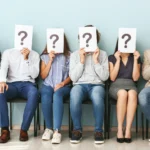 Gruppe von Bewerbenden sitzt im Wartebereich und hält Blätter mit Fragezeichen vor das Gesicht – zeigt Unsicherheit darüber, welche Faktoren bei der Jobwahl wirklich wichtig sind.