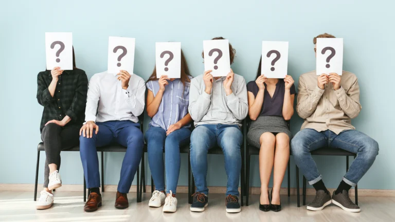 Gruppe von Bewerbenden sitzt im Wartebereich und hält Blätter mit Fragezeichen vor das Gesicht – zeigt Unsicherheit darüber, welche Faktoren bei der Jobwahl wirklich wichtig sind.