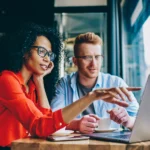 Zwei Menschen sitzen in einem Café vor einem Laptop, arbeiten gemeinsam und besprechen Inhalte. Das Bild vermittelt flexibles Arbeiten und zeigt ein modernes Arbeitsmodell mit freier Ortswahl außerhalb des Büros.