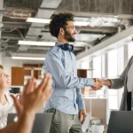 Team begrüßt einen neuen Mitarbeitenden im modernen Büro; eine Frau und ein Mann geben sich die Hand, während das Team applaudiert – erfolgreiche Integration über Arbeitnehmerüberlassung.