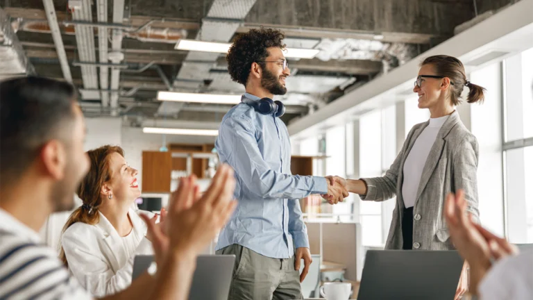 Team begrüßt einen neuen Mitarbeitenden im modernen Büro; eine Frau und ein Mann geben sich die Hand, während das Team applaudiert – erfolgreiche Integration über Arbeitnehmerüberlassung.