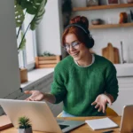 Junge Frau arbeitet im Homeoffice an einem Laptop, trägt Kopfhörer und lächelt während eines Online-Meetings – Beispiel für flexible Arbeitsmodelle und ortsunabhängiges Arbeiten.