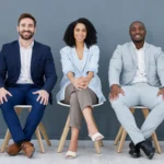 Fünf Personen in Business-Outfits sitzen nebeneinander auf Stühlen im Wartebereich eines Büros. Jede Person hält Bewerbungsunterlagen oder eine Mappe in den Händen.