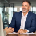 Berufserfahrener Bewerber im Gespräch mit einer Personalerin an einem Konferenztisch. Er trägt einen dunkelblauen Anzug, lächelt und wirkt professionell und selbstsicher. Modernes Büro im Hintergrund.