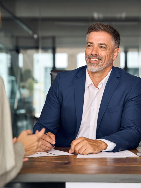 Berufserfahrener Bewerber im Gespräch mit einer Personalerin an einem Konferenztisch. Er trägt einen dunkelblauen Anzug, lächelt und wirkt professionell und selbstsicher. Modernes Büro im Hintergrund.