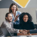 Drei Personen sitzen in einem modernen Büro bei einem Bewerbungsgespräch. Sie lächeln und geben einer vierten Person außerhalb des Bildausschnitts die Hand – ein Zeichen dafür, dass der Bewerbungsprozess erfolgreich abgeschlossen wurde.