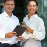 ChatGPT: Ein Mann und eine Frau in eleganter Business-Kleidung stehen vor einem modernen Bürogebäude und lächeln in die Kamera. Beide halten digitale Geräte in der Hand. Ein Beispiel für ein gepflegtes, professionelles Auftreten beim Vorstellungsgespräch.