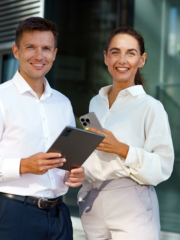 ChatGPT: Ein Mann und eine Frau in eleganter Business-Kleidung stehen vor einem modernen Bürogebäude und lächeln in die Kamera. Beide halten digitale Geräte in der Hand. Ein Beispiel für ein gepflegtes, professionelles Auftreten beim Vorstellungsgespräch.