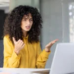 Eine Frau sitzt in einem modernen Büro vor einem Laptop und gestikuliert beim Sprechen, als würde sie ein Problem erklären oder Unzufriedenheit im Arbeitsalltag äußern.