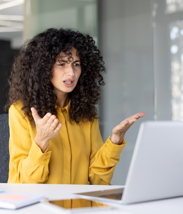Eine Frau sitzt in einem modernen Büro vor einem Laptop und gestikuliert beim Sprechen, als würde sie ein Problem erklären oder Unzufriedenheit im Arbeitsalltag äußern.