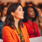 Junge Frau in einem orangefarbenen Blazer sitzt aufmerksam in einem Seminarraum; ihr Outfit kombiniert moderne Modeelemente mit einem professionellen Look, während andere Teilnehmende im Hintergrund ebenfalls business-lässige Kleidung tragen.