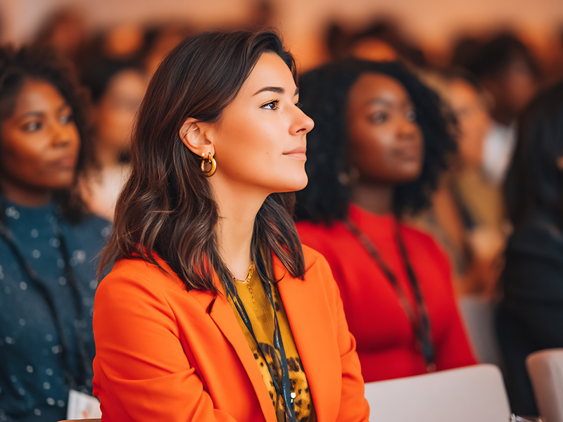 Junge Frau in einem orangefarbenen Blazer sitzt aufmerksam in einem Seminarraum; ihr Outfit kombiniert moderne Modeelemente mit einem professionellen Look, während andere Teilnehmende im Hintergrund ebenfalls business-lässige Kleidung tragen.