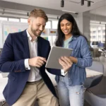 Ein Mann im Sakko und eine Frau im Jeanshemd stehen in einem modernen Büro und sehen sich gemeinsam Inhalte auf einem Tablet an. Beide lächeln, während sie über ein Projekt im Rahmen der Arbeitnehmerüberlassung sprechen.