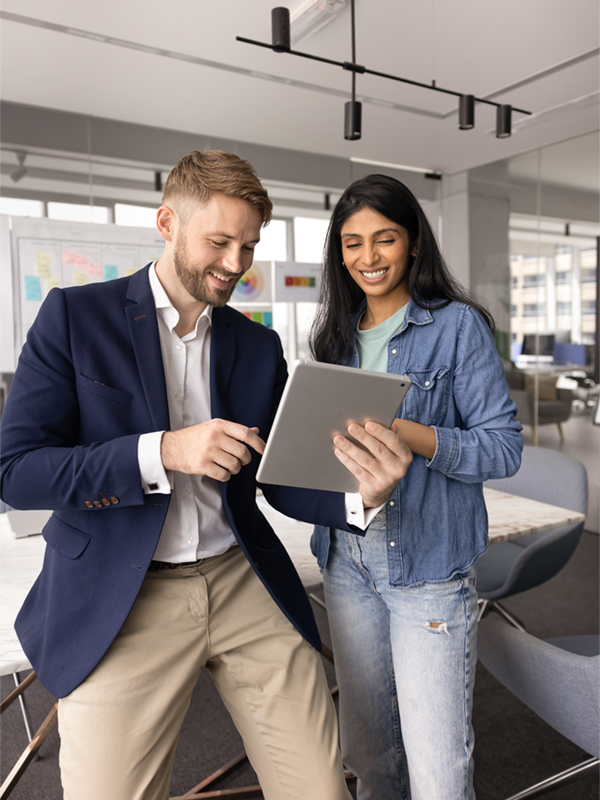 Ein Mann im Sakko und eine Frau im Jeanshemd stehen in einem modernen Büro und sehen sich gemeinsam Inhalte auf einem Tablet an. Beide lächeln, während sie über ein Projekt im Rahmen der Arbeitnehmerüberlassung sprechen.