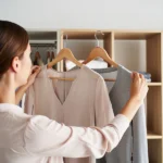 Eine Frau steht vor einem offenen Kleiderschrank und hält zwei Outfits, eine helle Bluse und einen grauen Pullover, nebeneinander, um das passende Kleidungsstück für ein Vorstellungsgespräch auszuwählen.