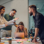 Vier Personen in einem modernen Büro diskutieren angeregt um einen Tisch mit Laptop und Notizen. Eine Frau sitzt mit den Händen an den Schläfen vor dem Bildschirm, während die anderen drei gestikulierend um sie herumstehen.
