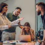 Vier Kolleg:innen stehen in einem Meetingraum und diskutieren lautstark, während eine Frau am Laptop sitzt und gestresst den Kopf hält – eine Situation, die zeigt, wie belastend ein unausgeglichenes Arbeitsumfeld sein kann.