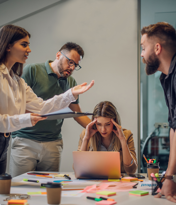 Vier Kolleg:innen stehen in einem Meetingraum und diskutieren lautstark, während eine Frau am Laptop sitzt und gestresst den Kopf hält – eine Situation, die zeigt, wie belastend ein unausgeglichenes Arbeitsumfeld sein kann.