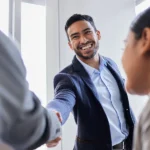 Ein Mann im blauen Sakko lächelt freundlich und reicht einer anderen Person im Büro die Hand zur Begrüßung. Eine Szene, die den erfolgreichen Start in ein neues Projekt oder eine Zusammenarbeit im Rahmen der Arbeitnehmerüberlassung zeigt.