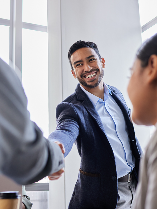 Ein Mann im blauen Sakko lächelt freundlich und reicht einer anderen Person im Büro die Hand zur Begrüßung. Eine Szene, die den erfolgreichen Start in ein neues Projekt oder eine Zusammenarbeit im Rahmen der Arbeitnehmerüberlassung zeigt.