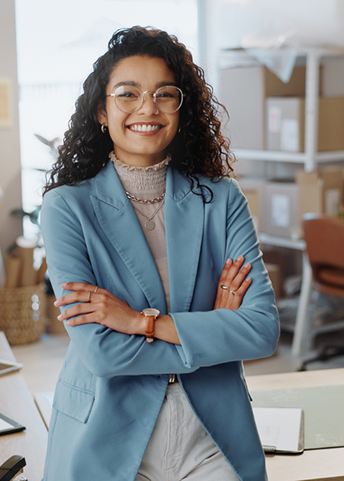 Junge Frau mit lockigem Haar und Brille steht lächelnd in einem Büro. Sie trägt einen hellblauen Blazer und eine helle Bluse – ein moderner, gepflegter Look, der Professionalität ausstrahlt.