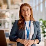 Frau mit rotbraunen Haaren steht in einem modernen, lichtdurchfluteten Büro neben einem Laptop. Sie trägt ein Namensband und lächelt freundlich in die Kamera. Im Hintergrund sind Pflanzen und Glaswände zu sehen.