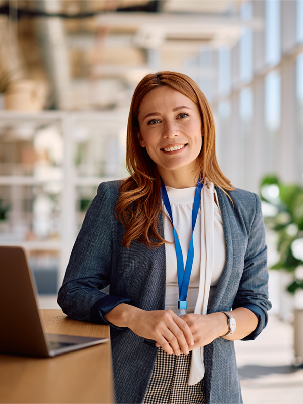 Frau mit rotbraunen Haaren steht in einem modernen, lichtdurchfluteten Büro neben einem Laptop. Sie trägt ein Namensband und lächelt freundlich in die Kamera. Im Hintergrund sind Pflanzen und Glaswände zu sehen.
