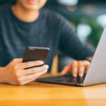 Person sitzt an einem Holztisch und nutzt gleichzeitig Laptop und Smartphone zur Online-Jobsuche auf einem Karriereportal.