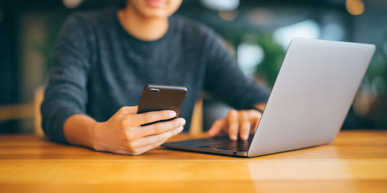 Person sitzt an einem Holztisch und nutzt gleichzeitig Laptop und Smartphone zur Online-Jobsuche auf einem Karriereportal.