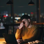 Ein Mann sitzt spätabends erschöpft und leicht verzweifelt vor einem Laptop, während er sich mit einer Hand die Augen reibt. Er trägt einen senfgelben Pullover, vor ihm liegen ein Notizbuch und ein Stift. Das Licht der Schreibtischlampe fällt warm auf sein Gesicht, während im Hintergrund die Lichter der Stadt durch das Fenster leuchten – er wirkt, als suche er frustriert nach passenden Jobs in Karriereportalen.