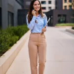 Junge Frau in einem business-casual Outfit für ein Vorstellungsgespräch, bestehend aus einer hellblauen Bluse, beigefarbenen Stoffhosen und braunen Loafern. Sie steht im Freien vor modernen Bürogebäuden, lächelt und telefoniert, während sie ein Tablet in der Hand hält. Das Outfit wirkt professionell, aber zugleich modern und nahbar – ideal für ein Bewerbungsgespräch im Büro- oder Dienstleistungsumfeld.