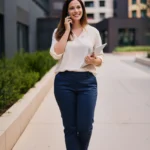Frau in einem Business-Casual-Outfit für ein Vorstellungsgespräch, bestehend aus einer cremefarbenen Bluse, dunkelblauer Stoffhose und schwarzen Loafern. Sie steht vor modernen Bürogebäuden, lächelt und telefoniert, während sie ein Tablet in der Hand hält. Das Outfit vermittelt einen gepflegten, professionellen und dennoch entspannten Eindruck – ideal für Bewerbungsgespräche in Unternehmen mit moderatem Dresscode.