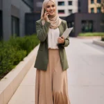 Frau in einem Business-Casual-Outfit für ein Vorstellungsgespräch, bestehend aus einem olivgrünen Blazer, einer cremefarbenen Bluse, einem beigen Maxirock und einem farblich passenden Hijab. Sie trägt schwarze Flats und hält ein Tablet in der Hand, während sie lächelnd telefoniert. Das Outfit wirkt professionell, elegant und zugleich modern – ideal für Bewerbungsgespräche mit Fokus auf Stil und Seriosität.